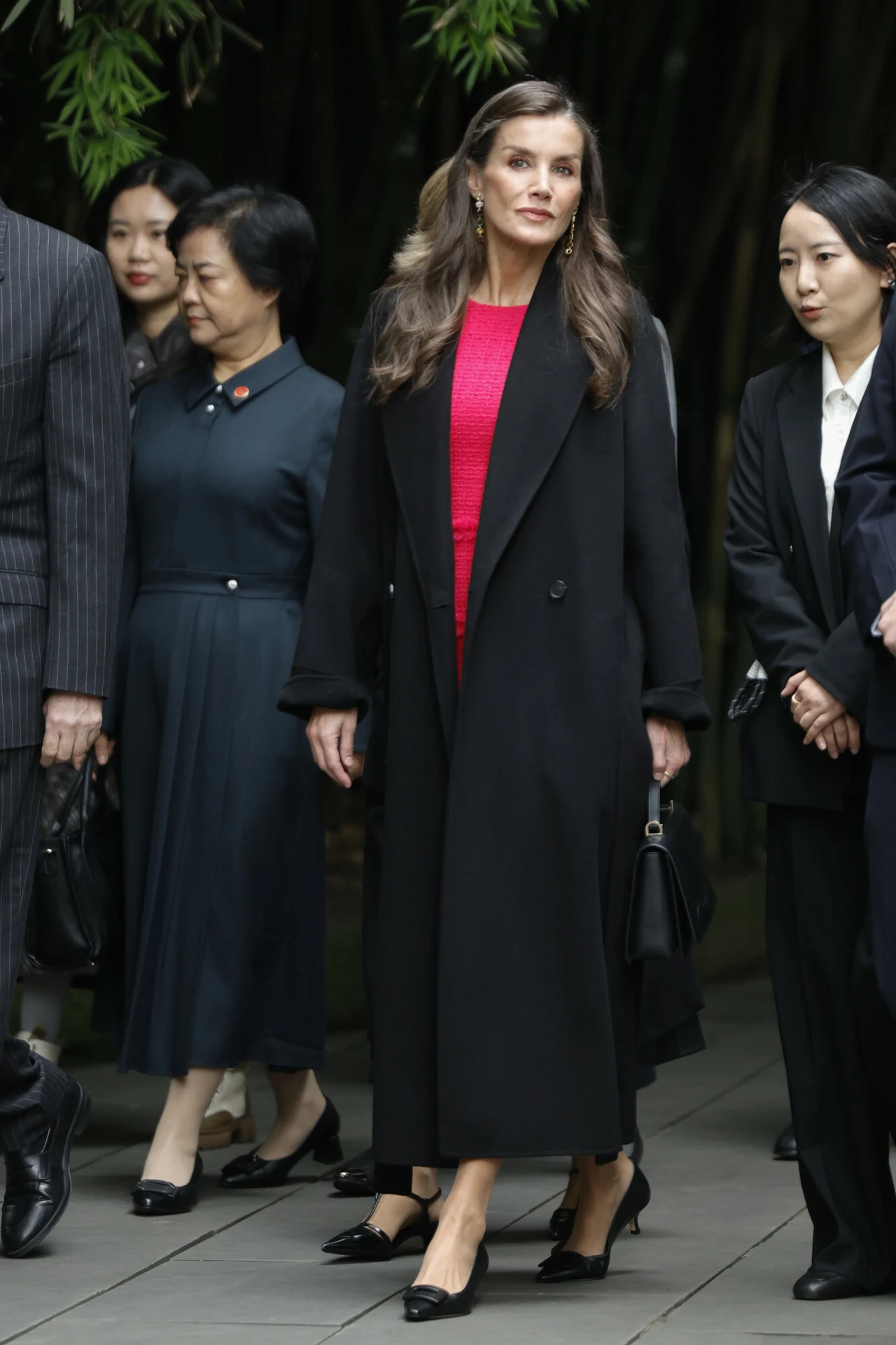 Grupa elegancko ubranych osób przemieszczających się wspólnie, w centrum kobieta w długim czarnym płaszczu i czerwonej sukni, której wygląd wyraźnie przyciąga uwagę. W tle widać kilka innych osób, ubrane w ciemne stroje formalne.