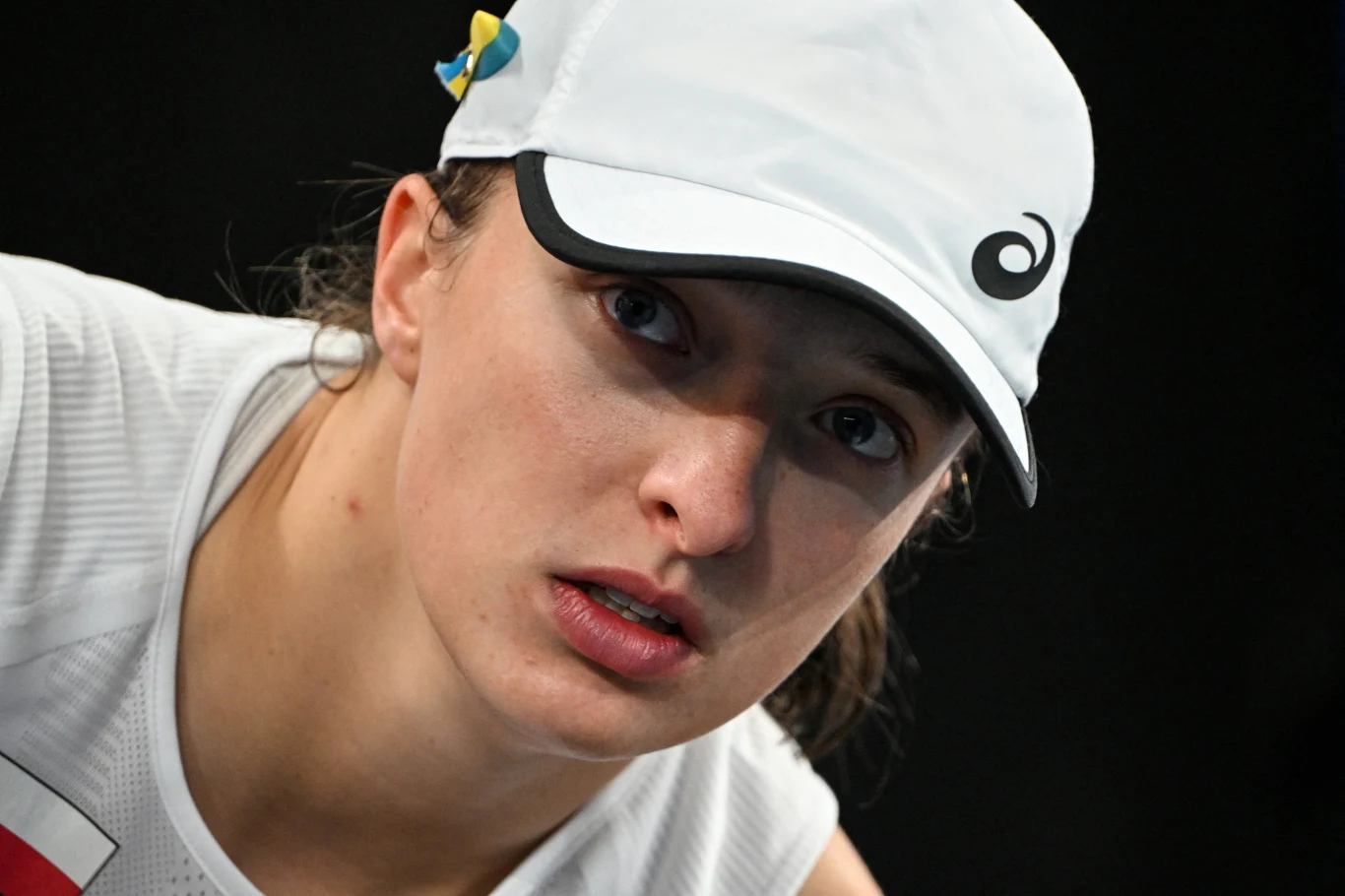 Iga Świątek Kobieta w białej czapce sportowej i białej koszulce, z intensywnym spojrzeniem, widoczna z bliska na czarnym tle, na czapce widoczne kolorowe ozdoby.