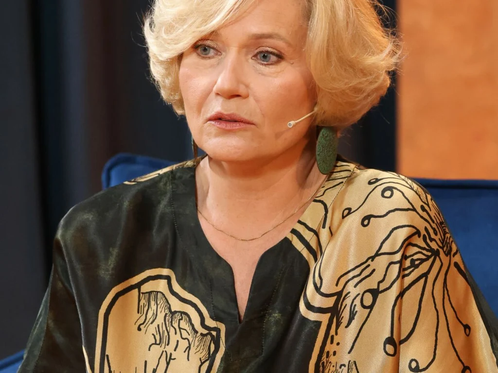 Kobieta o blond włosach w eleganckiej, wzorzystej bluzce siedzi na niebieskim fotelu, ma założony mikrofon na policzku i spogląda w bok z poważnym wyrazem twarzy.