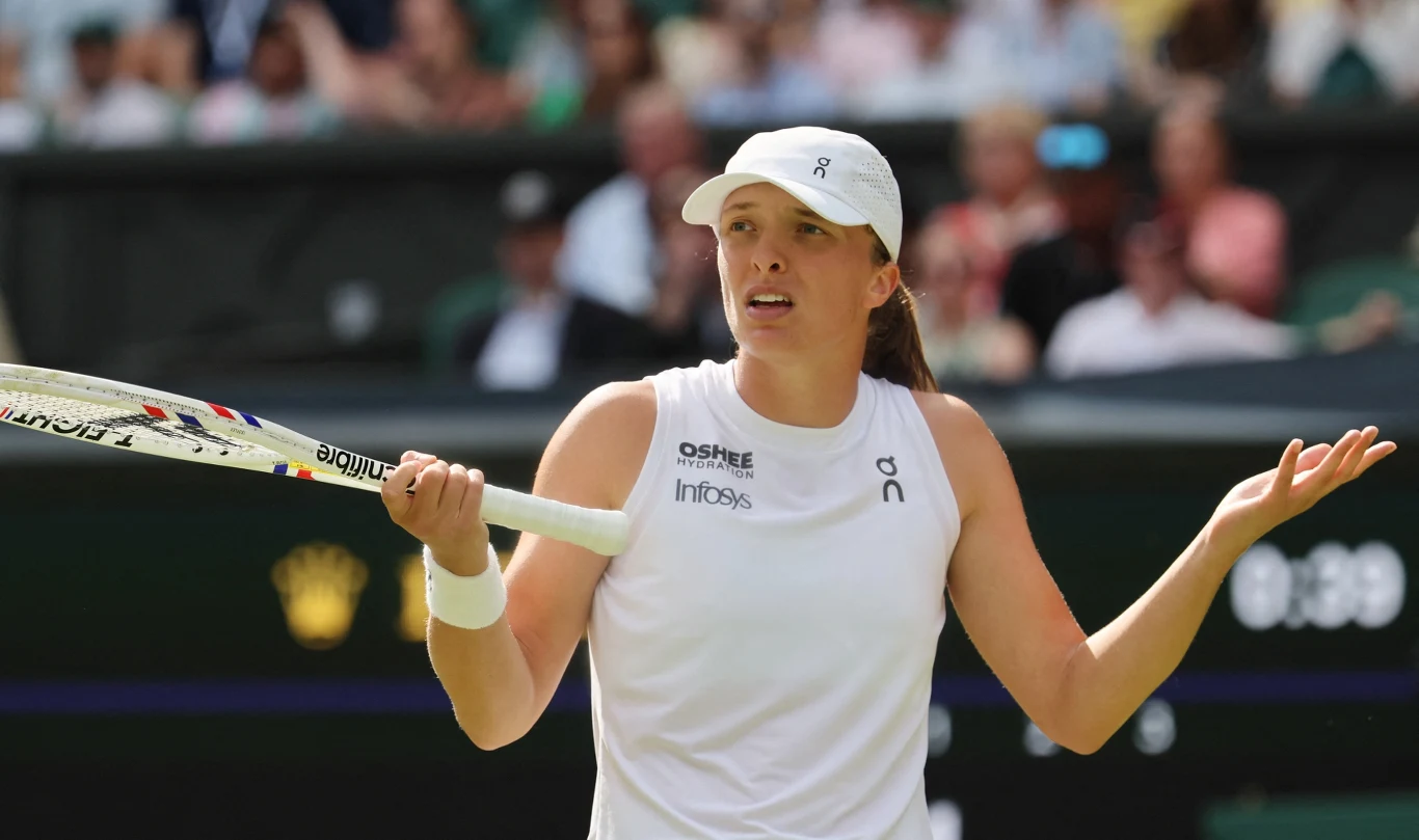 Tenisistka w białym stroju trzymająca rakietę, z wyrazem zdziwienia lub zaskoczenia na twarzy, na tle rozmytej publiczności.