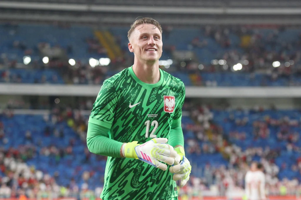 Marcin Bułka Bramkarz w zielonym stroju z numerem 12 i herbem Polski na piersi stoi na stadionie, w tle widoczna rozmyta publiczność na trybunach.