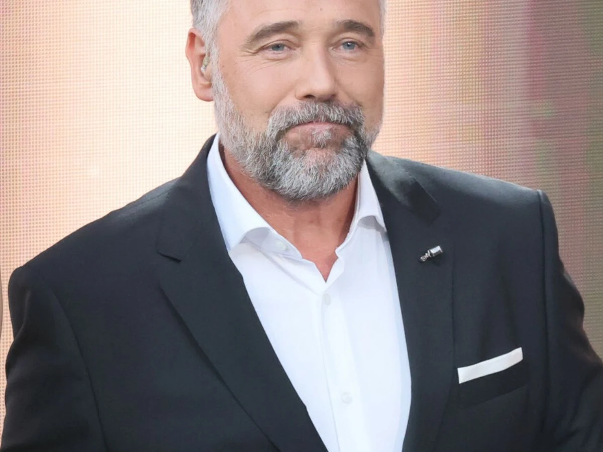 Łukasz Nowicki Mężczyzna w średnim wieku z siwą brodą i krótkimi włosami ubrany w ciemny garnitur oraz białą koszulę, z lekkim uśmiechem, stoi na tle jasnej, rozmytej scenerii.