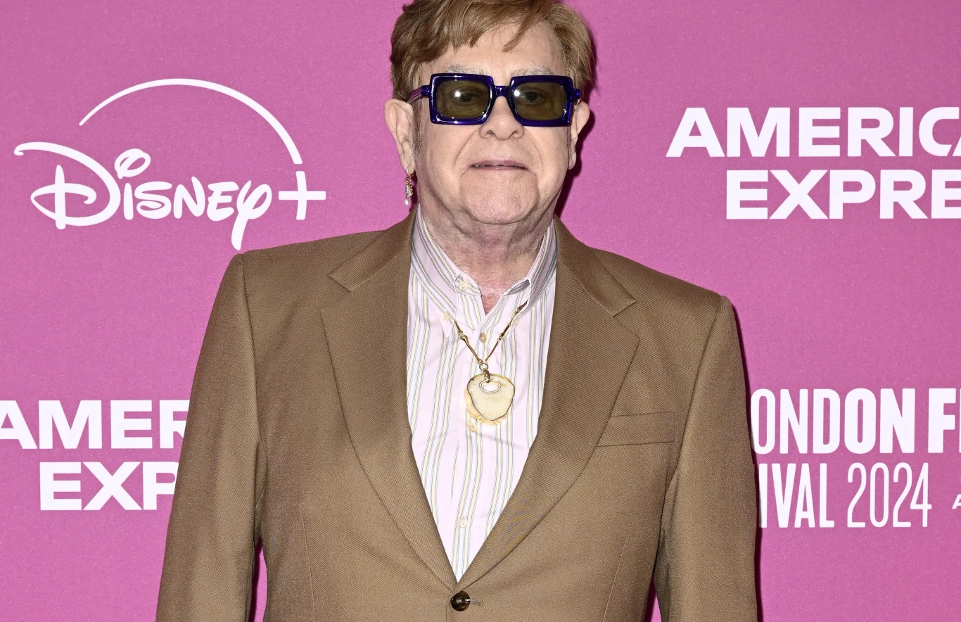 Elton John Mężczyzna w brązowej marynarce i pasiastą koszulą pod spodem, noszący ciemne okulary i biżuterię na szyi, stoi na tle fioletowej ścianki z logotypami Disney+ i American Express.
