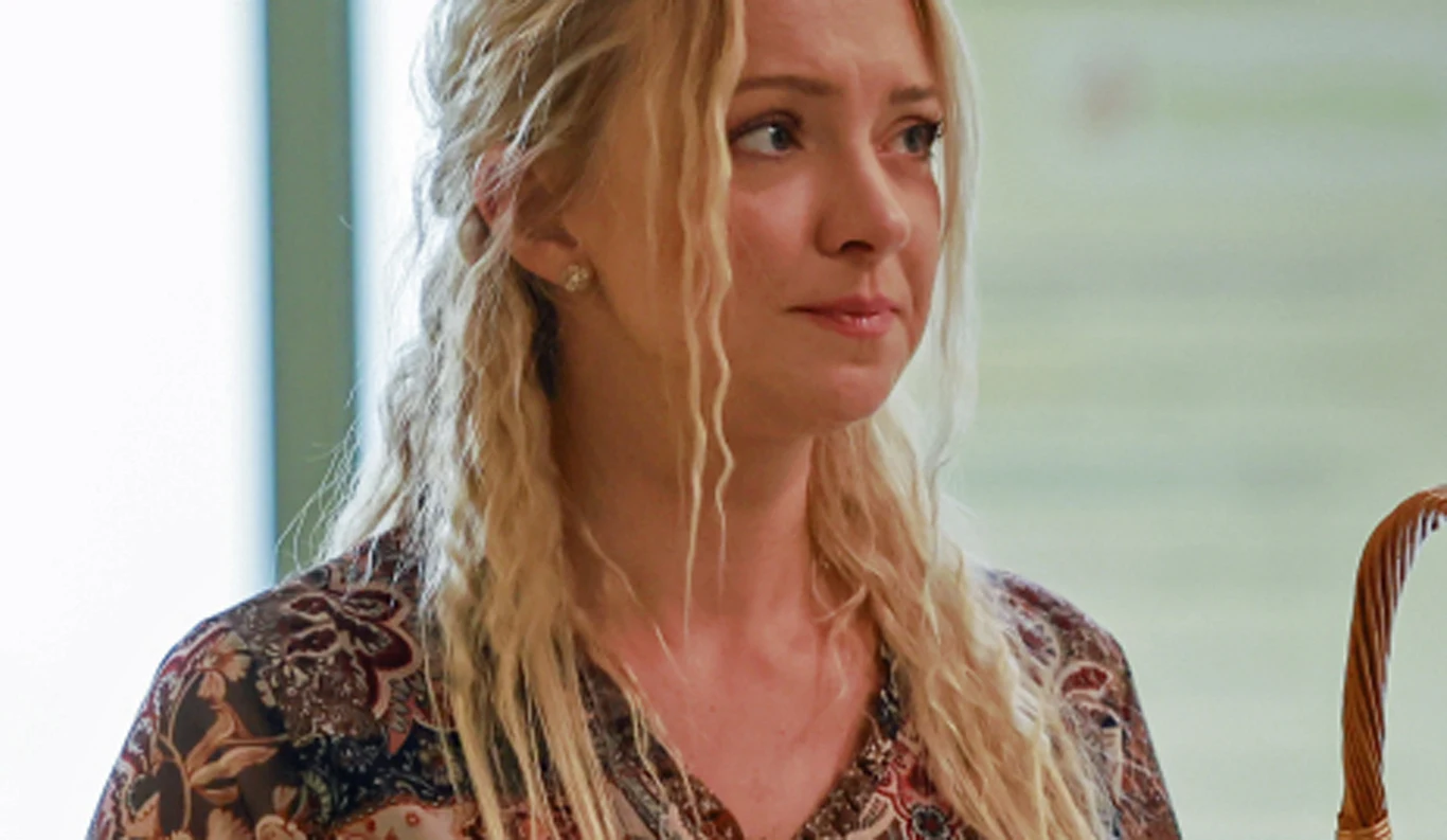 Barbara Kurdej-Szatan Blondynka o długich, falowanych włosach, ubrana w wzorzystą bluzkę, patrzy lekko w bok z wyrazem skupienia na twarzy. W tle widoczne rozmyte wnętrze pomieszczenia.