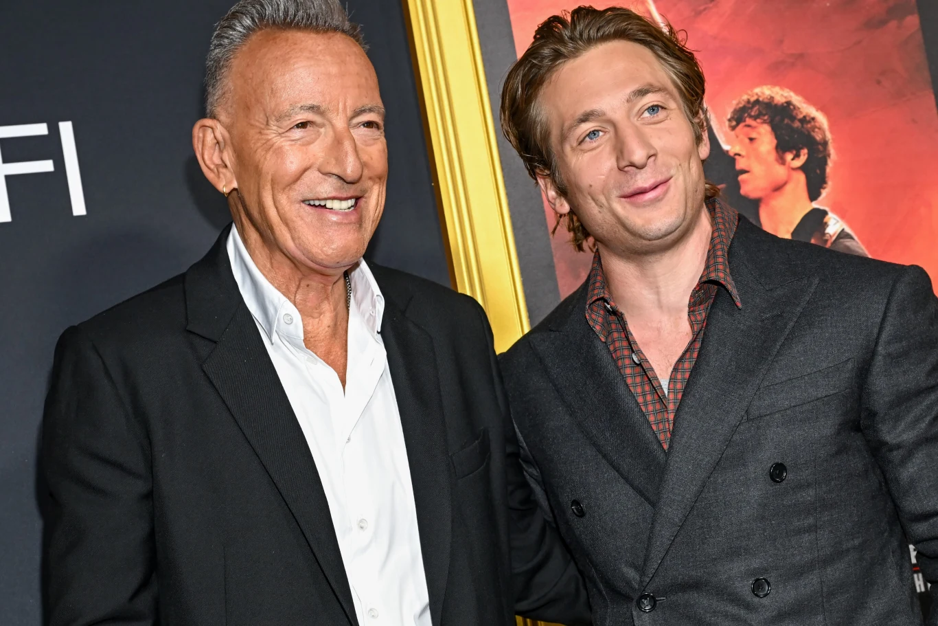 Bruce Springsteen i Jeremy Allen White promując film "Springsteen: Ocal mnie od nicości" Dwóch uśmiechniętych mężczyzn w eleganckich garniturach stoi razem na tle plakatu filmowego, jeden z nich trzyma drugiego za ramię.