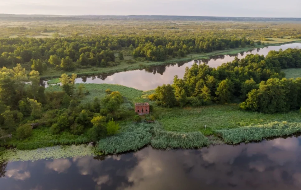 Prezydent Karol Nawrocki zawetował ustawę o utworzeniu Parku Narodowego Doliny Dolnej Odry / fot. Piotr Piznal / Ministerstwo Klimatu i Środowiska Zabytkowy budynek z cegły otoczony gęstą zielenią nad brzegiem spokojnej rzeki, w tle rozległe tereny podmokłe i lasy sięgające aż po horyzont.