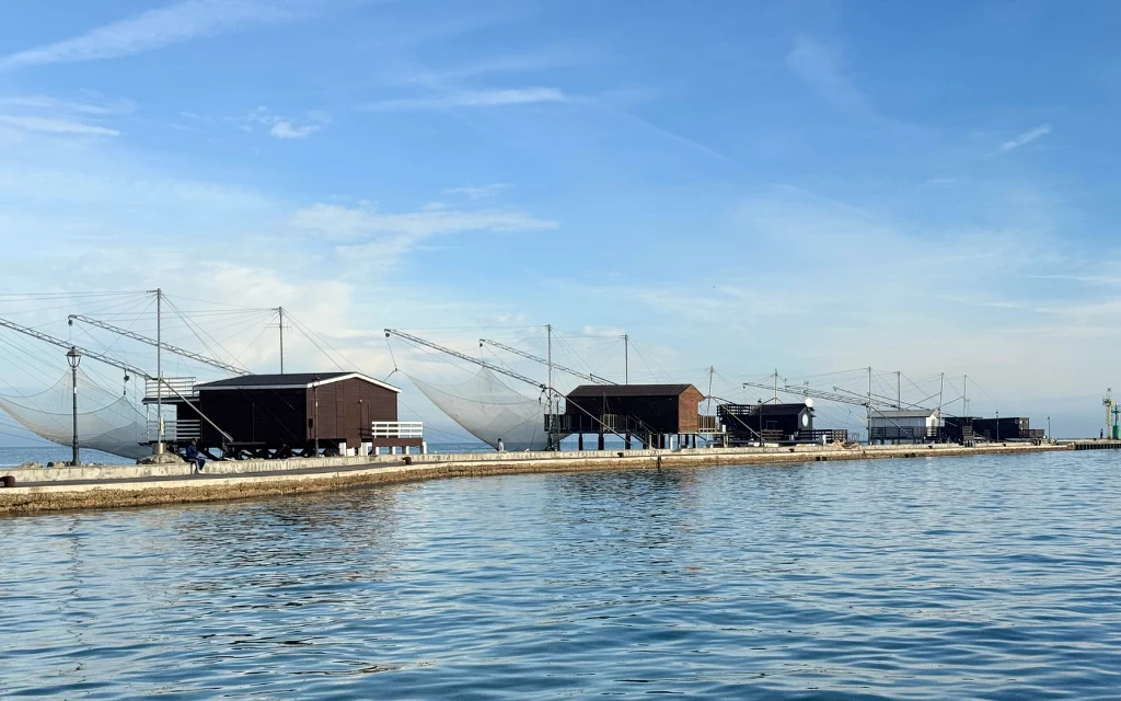 Cesenatico oddycha morzem Szereg drewnianych domków rybackich wzdłuż brzegu z dużymi sieciami rozpiętymi na masztach nad spokojną wodą, błękitne niebo z lekkimi chmurami.