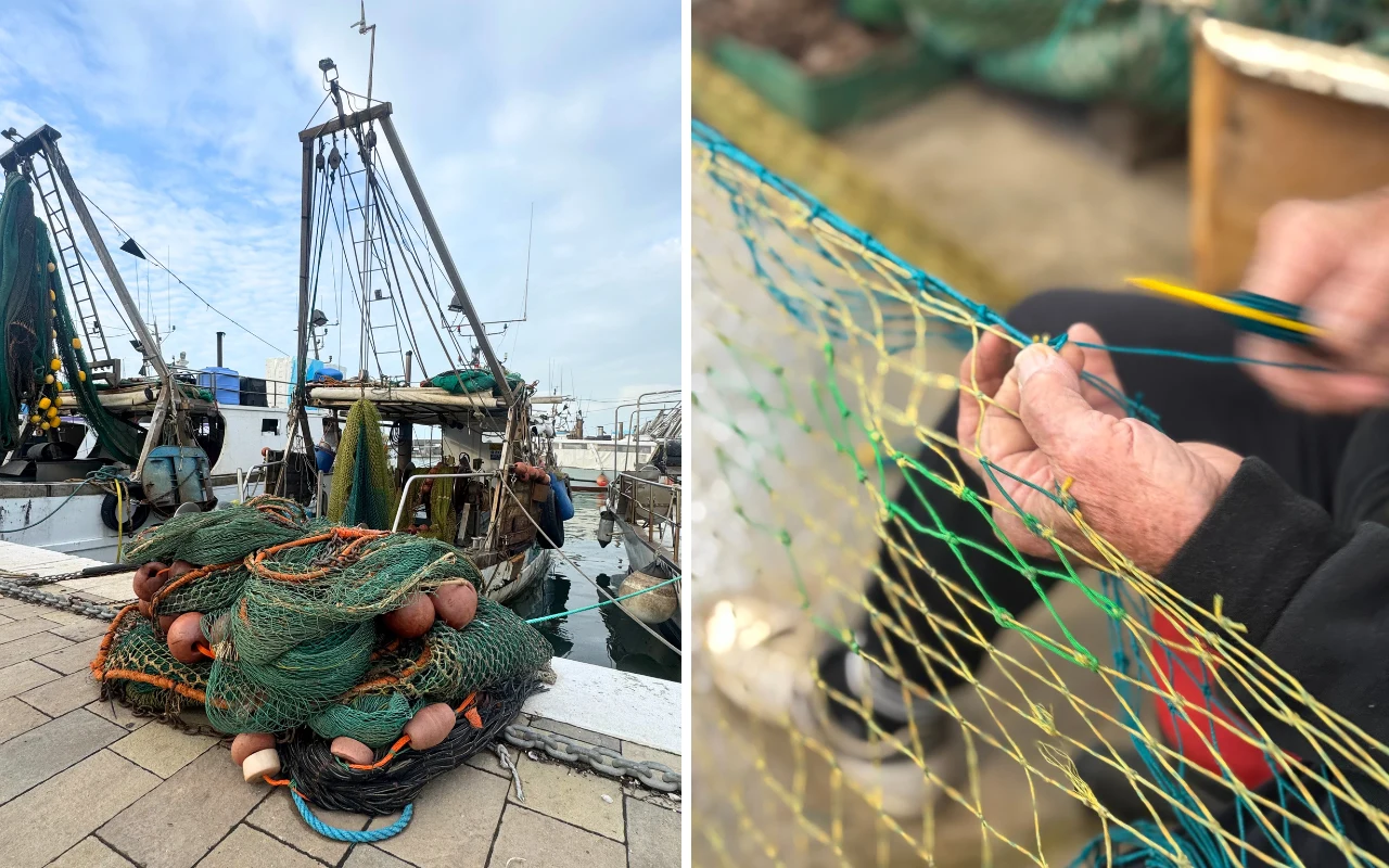 Stosy zielonych i pomarańczowych sieci rybackich leżą na nabrzeżu portowym, w tle cumują kutry rybackie. Po prawej stronie widoczne są dłonie osoby naprawiającej sieć rybacką grubą igłą.