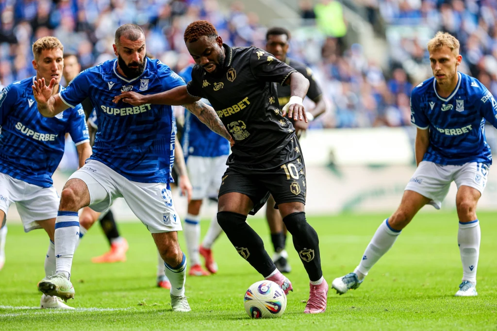 Mikael Ishak i Afimico Pululu w walce o piłkę Zawodnicy drużyn piłkarskich w niebieskich i czarnych strojach walczą o piłkę podczas meczu na stadionie, wokół nich widać skupionych rywali i trybuny z kibicami w tle.