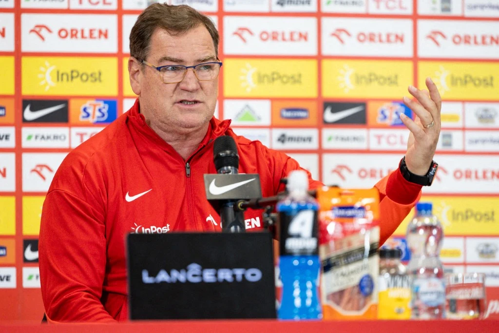 Jan Urban przed meczem Holandia - Polska Mężczyzna w czerwonym dresie siedzi przy stole podczas konferencji prasowej, przemawia do mikrofonu, w tle widoczne są logotypy sponsorów oraz napoje sportowe i butelka wody na stole.