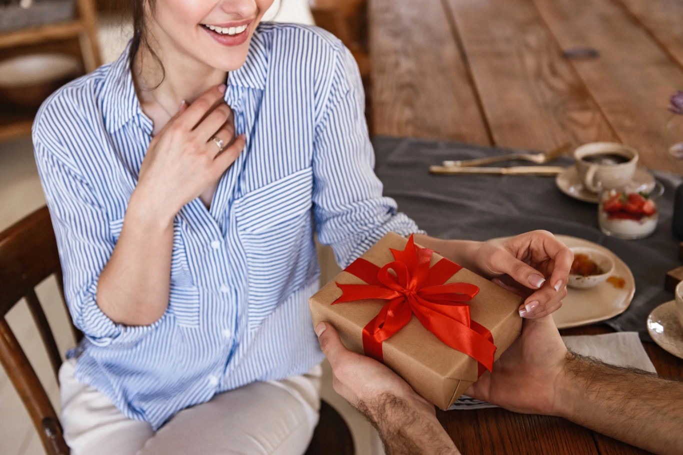 Perfumy idealnie nadają się na prezent dla bliskiej osoby Kobieta w jasnej koszuli z uśmiechem przyjmuje prezent owinięty w brązowy papier i ozdobiony czerwoną wstążką, siedzi przy drewnianym stole na którym stoją filiżanki i talerzyki.