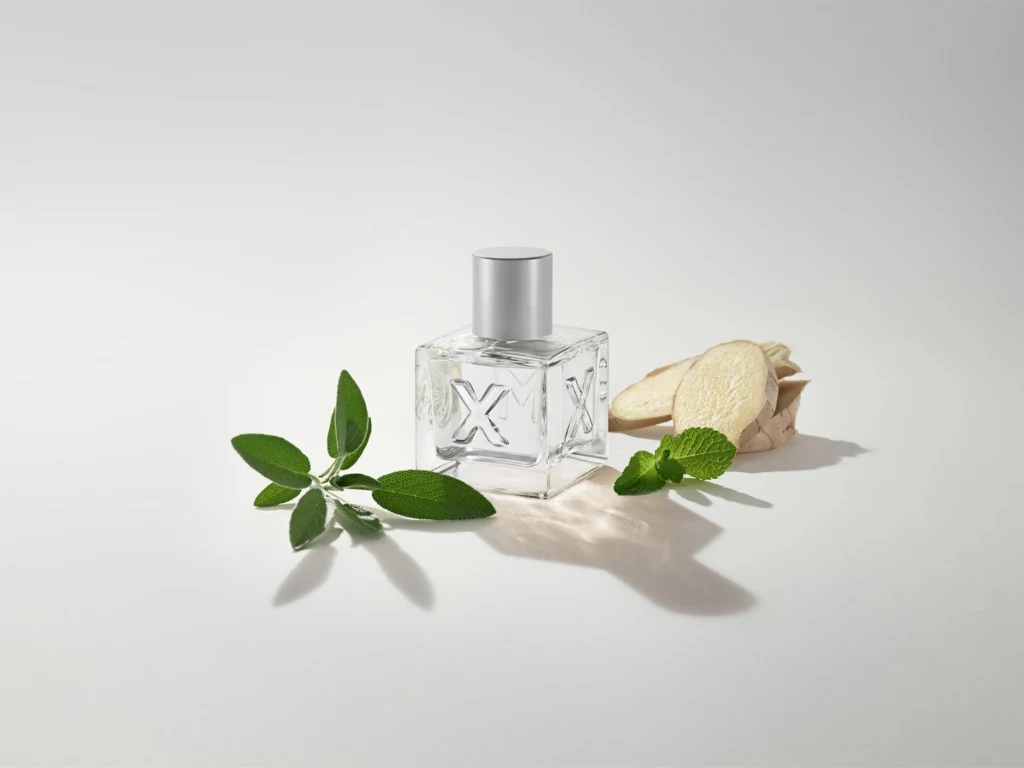 Kwiatowo-waniliowy zapach z nutami crème brûlée i brzoskwini kusi zmysłowością Butelka perfum o przezroczystych ściankach i srebrnym korku, umieszczona obok świeżo ściętych liści ziołowych i kawałków korzenia imbiru, całość utrzymana na jasnym, minimalistycznym tle.