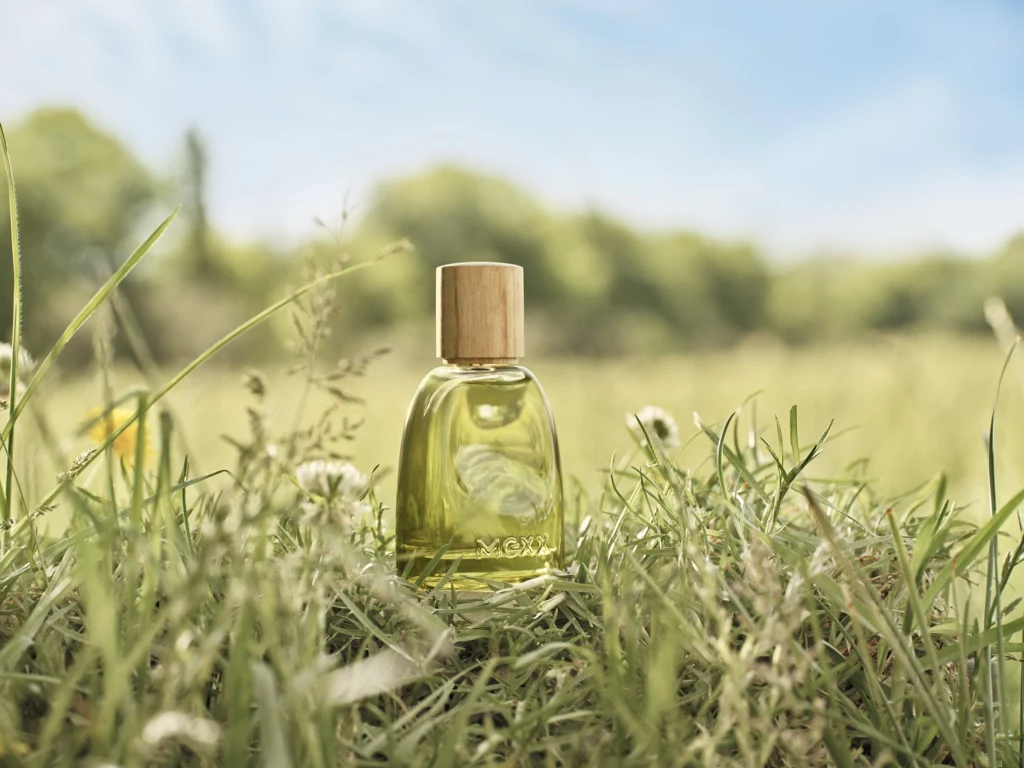 Kompozycja MEXX Inspired Nature dla niej i dla niego otula znaki ziemskie kwiatowo-owocowymi nutami Butelka perfum z drewnianą nakrętką ustawiona pośród zielonej trawy na tle rozmytej, słonecznej łąki i błękitnego nieba.