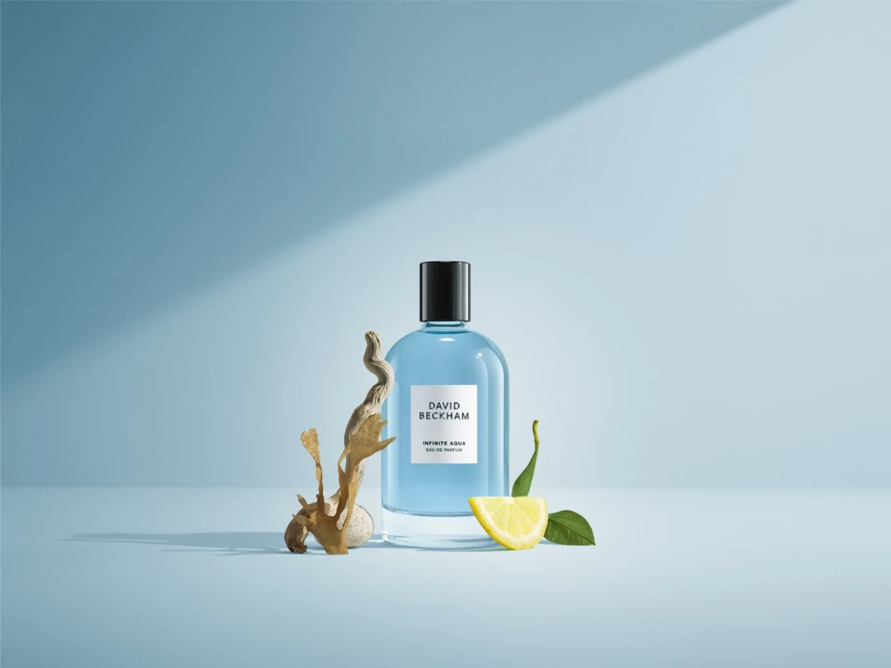 Znaki wodne cenią sobie elegancję i ponadczasowy styl Butelka perfum sygnowana marką David Beckham ustawiona na niebieskim tle, obok której znajduje się plasterek cytryny z liściem oraz element korzenia, wszystko oświetlone miękkim światłem padającym z prawej strony.