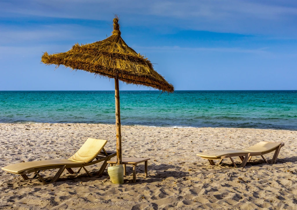 Plaża piaszczysta z parasolem z trzciny oraz dwoma leżakami skierowanymi w stronę błękitnego morza, spokojna tafla wody pod bezchmurnym niebem.