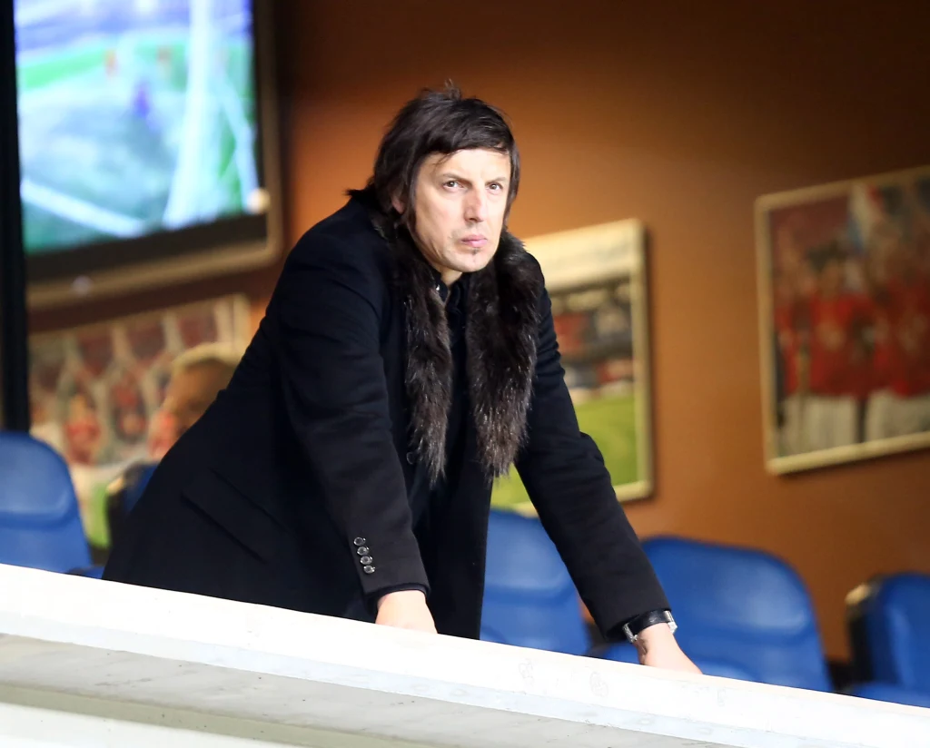 Mężczyzna w ciemnym płaszczu z futrzanym kołnierzem stoi oparty o balustradę na stadionie, za nim widoczne są niebieskie siedzenia i rozmyte plakaty piłkarskie na ścianie.