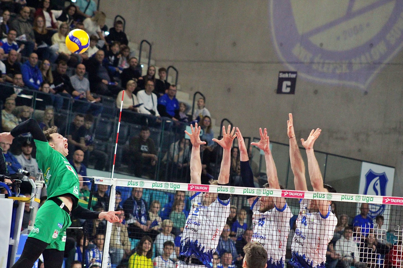 Kadr z meczu Ślepsk Malow Suwałki - Indykpol AZS Olsztyn Zawodnik w zielonym stroju wykonuje atak siatkarski nad siatką, blokowany przez trzech rywali w biało-niebieskich koszulkach, a w tle znajduje się licznie zgromadzona publiczność.