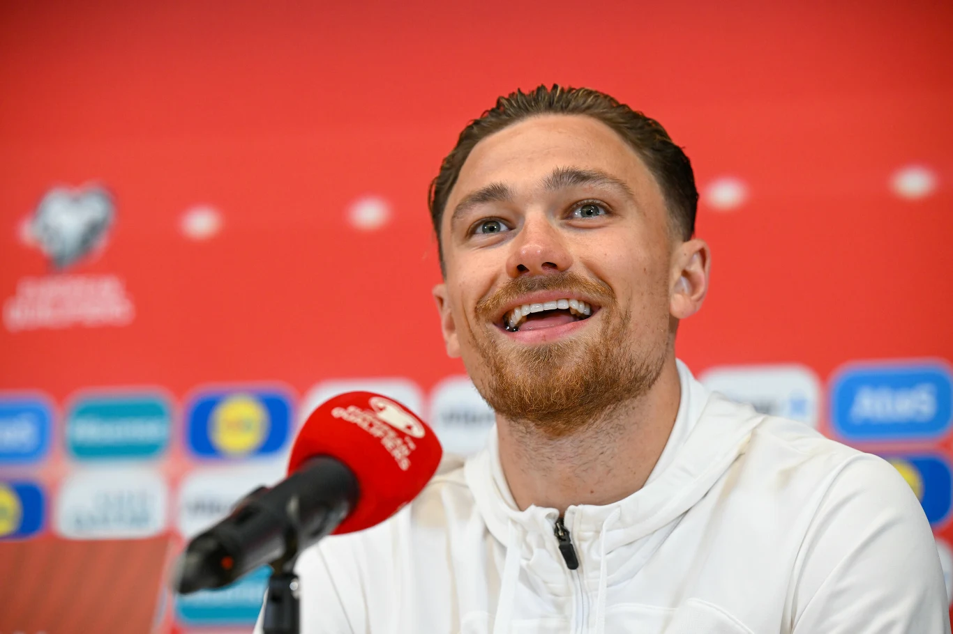 Uśmiechnięty mężczyzna w białej bluzie rozmawia z mediami przez mikrofon na tle czerwonej ściany z logotypami sponsorów.