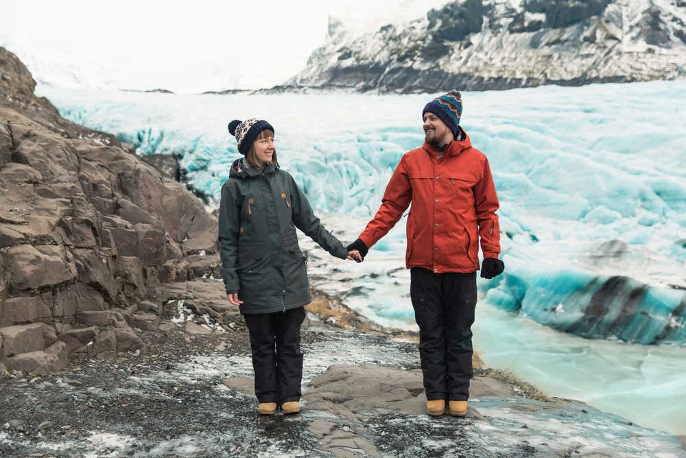 Para ubrana w zimowe kurtki i czapki stoi na skalistym terenie obok lodowca, trzymając się za ręce i patrząc na siebie. W tle widoczny masywny lodowiec oraz ośnieżone góry.