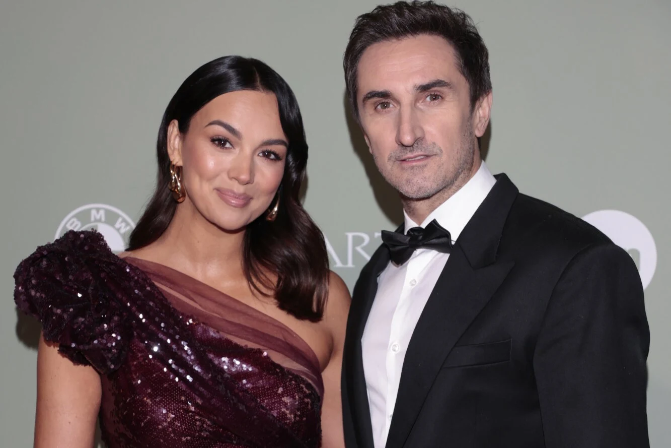 Paulina Krupińska-Karpiel i Sebastian Karpiel-Bułecka Elegancka para pozuje razem na tle jednolitej ściany. Kobieta ma na sobie ciemnofioletową, błyszczącą suknię z jednym odsłoniętym ramieniem, a mężczyzna ubrany jest w czarny smoking i muchę.