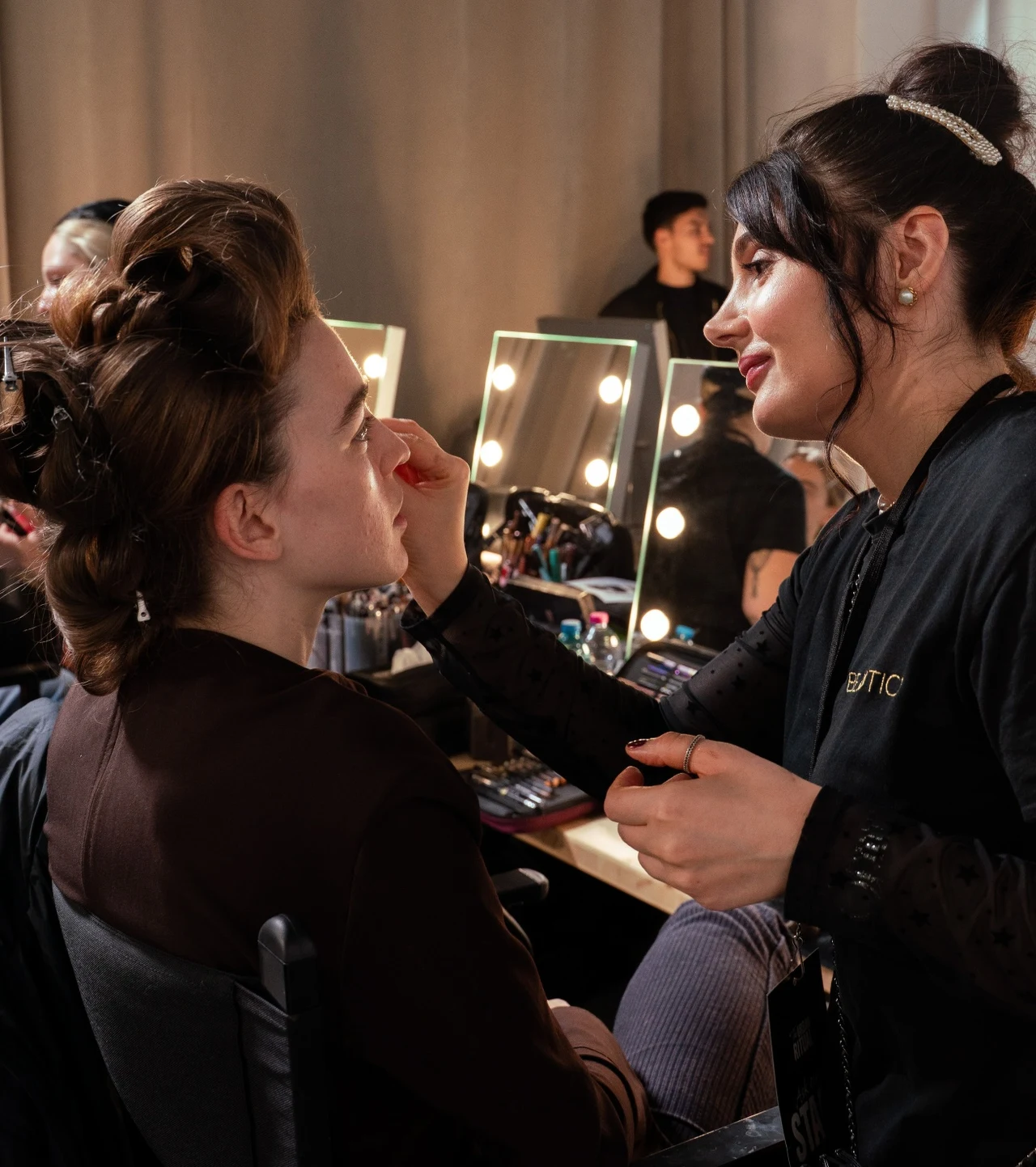 Pokaz SHEIN w Mediolanie: za makijaże modelek i modeli odpowiadała marka MIA Cosmetics – oficjalny partner pokazu Makijażystka wykonuje makijaż młodej kobiecie siedzącej na krześle w profesjonalnej garderobie. W tle lustra z oświetleniem i kosmetyki ułożone na stole, atmosfera skupienia i staranności.