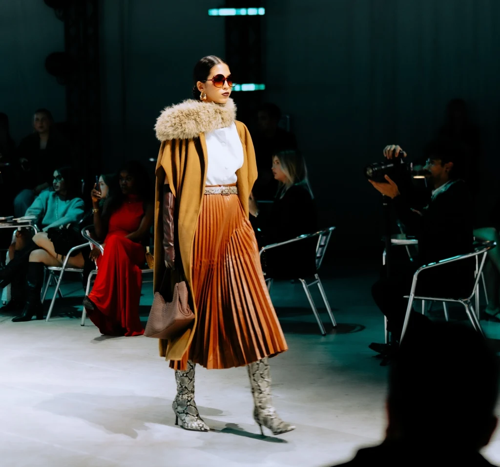Pokaz SHEIN w Mediolanie: pokaz w ramach projektu SHEIN X – inicjatywy wspierającej początkujących projektantów Modelka w brązowej plisowanej spódnicy, białej bluzce i płaszczu z futrzanym kołnierzem spaceruje po wybiegu. Ma ciemne okulary przeciwsłoneczne i wysokie buty w zwierzęcy wzór. W tle publiczność i fotograf.