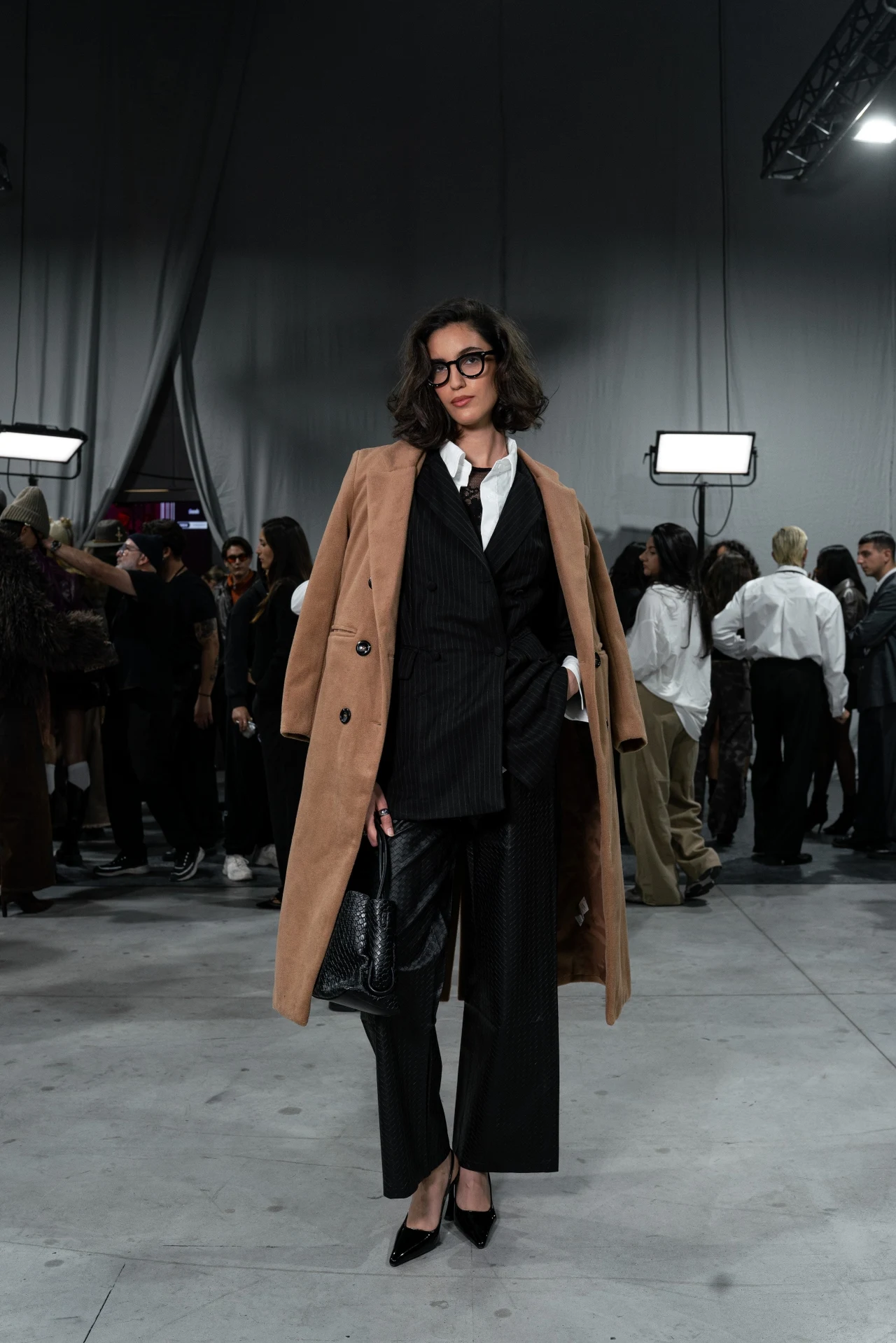 Pokaz SHEIN w Mediolanie: marka Missguided łącząca elegancję z nieszablonowym twistem Elegancko ubrana kobieta w okularach, czarnym garniturze, białej koszuli i jasnobrązowym płaszczu, trzymająca czarną torebkę, stoi na tle grupy ludzi w hali o industrialnym charakterze.