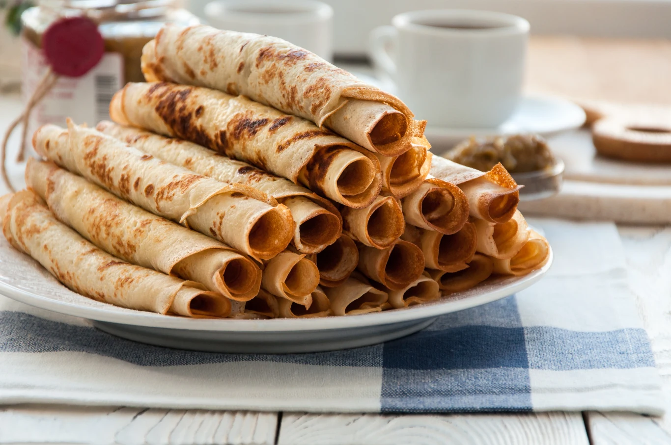 Stos cienkich naleśników zwiniętych w rulon ułożonych na białym talerzu, na tle zastawy z filiżankami i dodatkami na drewnianym stole.