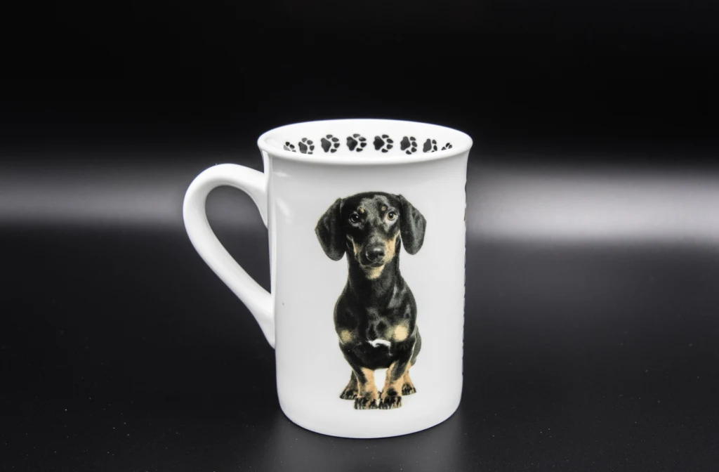Kubek z motywem jamnika? To tylko jedna z odsłon popularności tej rasy w obecnych czasach Biały kubek z wizerunkiem czarnego jamnika na przedniej części oraz śladami psich łapek wzdłuż górnej krawędzi wewnątrz naczynia, ustawiony na czarnym tle.