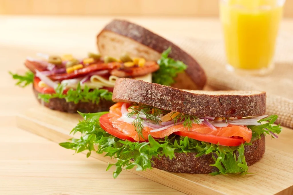 Dobry, ciemny chleb dodaje każdej kanapce smaku, zdrowia i charakteru Dwa kanapki na ciemnym chlebie z dużą ilością świeżej sałaty, pomidorem, cebulą, koperkiem i plastrem łososia lub innych dodatków, w tle szklanka świeżo wyciskanego soku pomarańczowego.