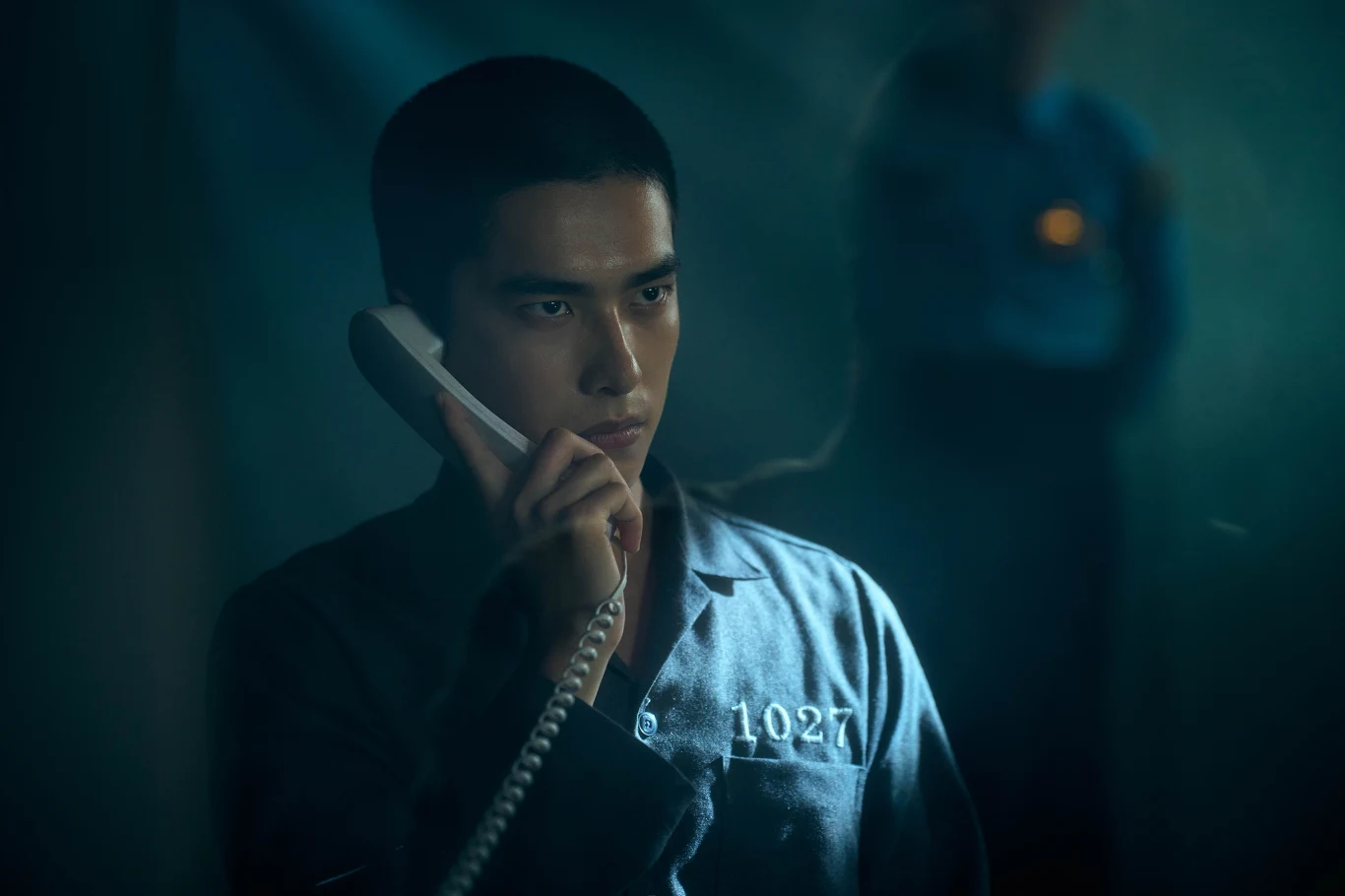 Młody mężczyzna w więziennej piżamie numer 1027 rozmawia przez telefon, z poważnym wyrazem twarzy, w tle widoczny nieostry strażnik.