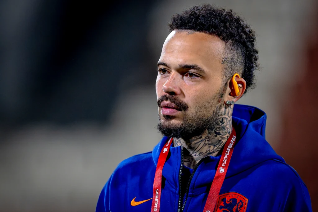 Mężczyzna z tatuażami na szyi, ubrany w niebieską kurtkę sportową z pomarańczowymi elementami, na szyi zawieszony identyfikator, w uchu żółta zatyczka. Tło rozmyte, skupienie na twarzy i mimice.