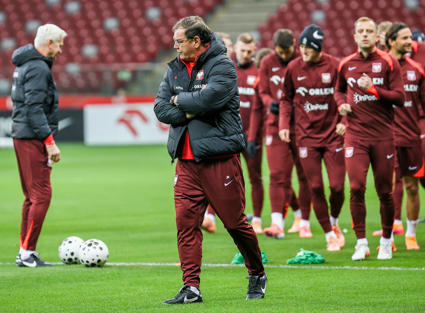 Grupa piłkarzy w stroju treningowym oraz trenerzy na murawie stadionu podczas sesji treningowej, piłki i akcesoria sportowe na trawie, trybuny w tle.