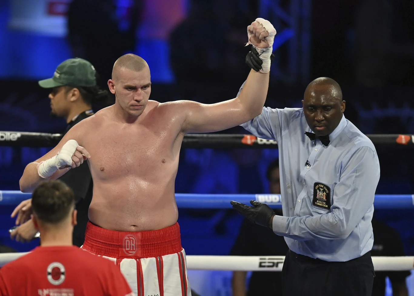 Zwycięski bokser stoi na ringu z uniesioną przez sędziego ręką, wyrażając zwycięstwo po walce. Bokser jest bez koszulki, w czerwonych spodenkach, sędzia ubrany jest w niebieską koszulę z emblematem. W tle widoczni są inni uczestnicy i nieostre sylwetki...