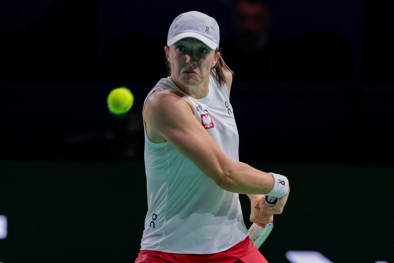 Iga Świątek podczas Billie Jean King Cup w barwach reprezentacji Polski Zawodniczka tenisa ziemnego w białej czapce i koszulce z polskim godłem, wykonująca uderzenie rakietą podczas meczu, na pierwszym planie żółta piłka tenisowa.