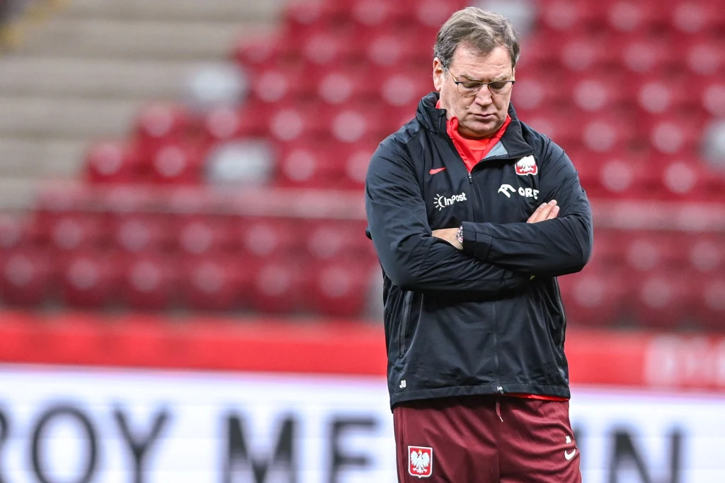 Mężczyzna w sportowej kurtce z logo Polski na piersi, stojący na stadionie z założonymi rękami, wyrażający zamyślenie lub rozczarowanie.