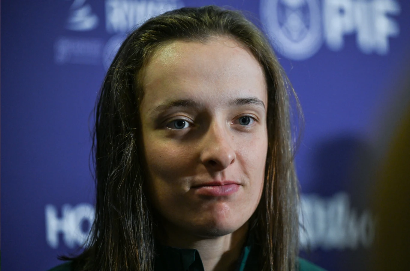 Młoda kobieta z długimi, ciemnymi włosami na fioletowym tle z niewyraźnymi logotypami. Twarz pokazana jest z bliska, wyraz subtelny i spokojny.