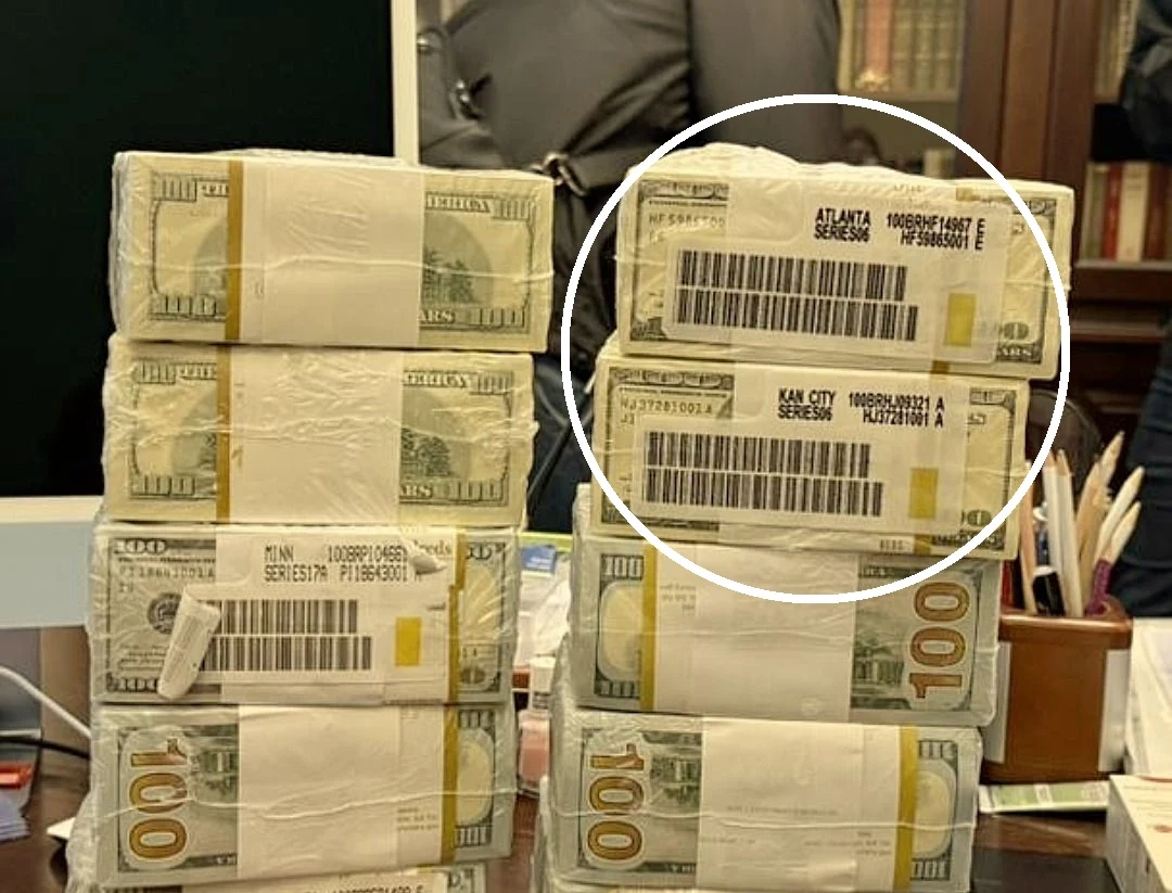 Zdjęcie znalezionych pieniędzy i amerykańskie etykiety Stosy banknotów stuzłotowych amerykańskiej waluty spięte białymi opaskami z kodami kreskowymi ułożone na biurku, w tle fragment sylwetki osoby i półka z książkami.