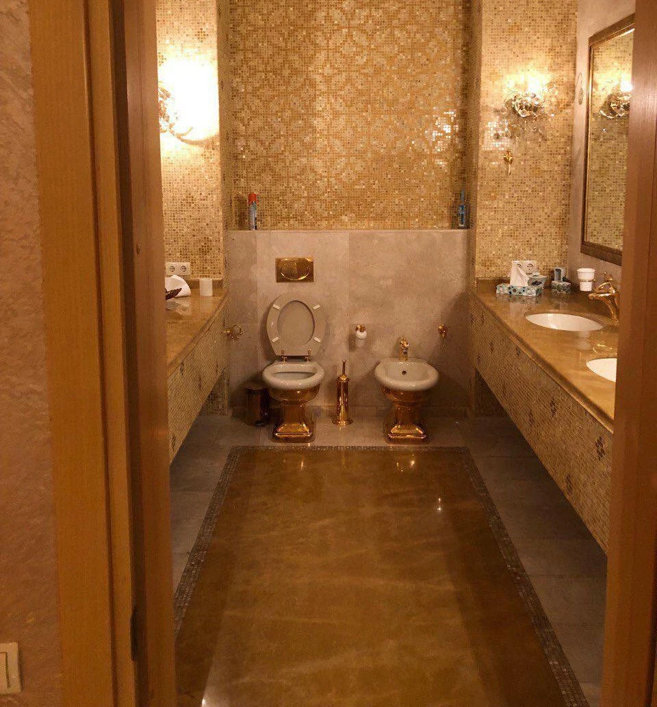 Luksusowa łazienka z bogato zdobionymi, złoconymi detalami. Dwa duże lustra, marmurowy blat z dwiema umywalkami, toaleta i bidet ozdobione złotymi akcentami. Ściany i podłogi pokryte dekoracyjną mozaiką oraz efektownym wzorem.