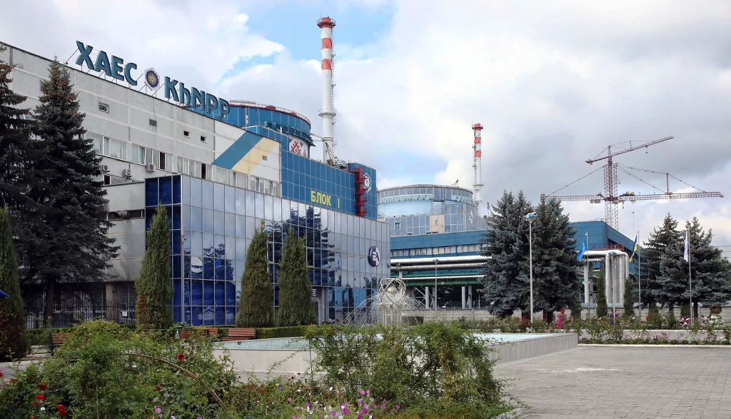 Kompleks budynków elektrowni jądrowej z widocznymi charakterystycznymi kominami i nowoczesną fasadą szklaną, otoczony zielenią i alejkami. Po prawej stronie żurawie budowlane, na pierwszym planie rabata kwiatowa i krzewy.