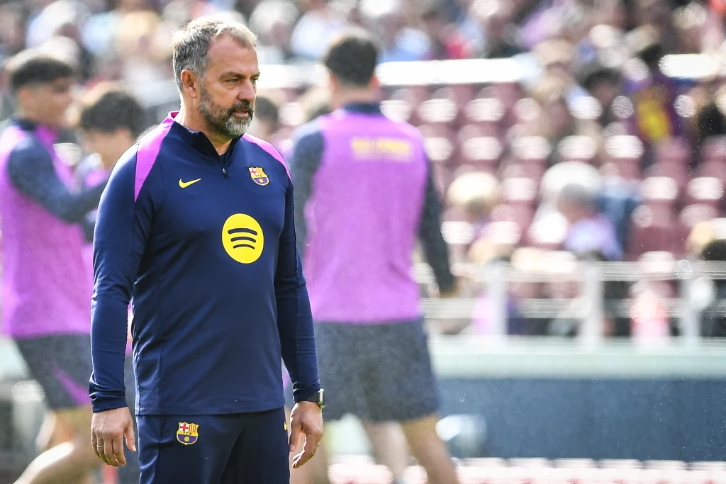 Trener w stroju FC Barcelona stoi na murawie stadionu, w tle widoczni są zawodnicy w fioletowych kamizelkach, trybuny wypełnione są kibicami.