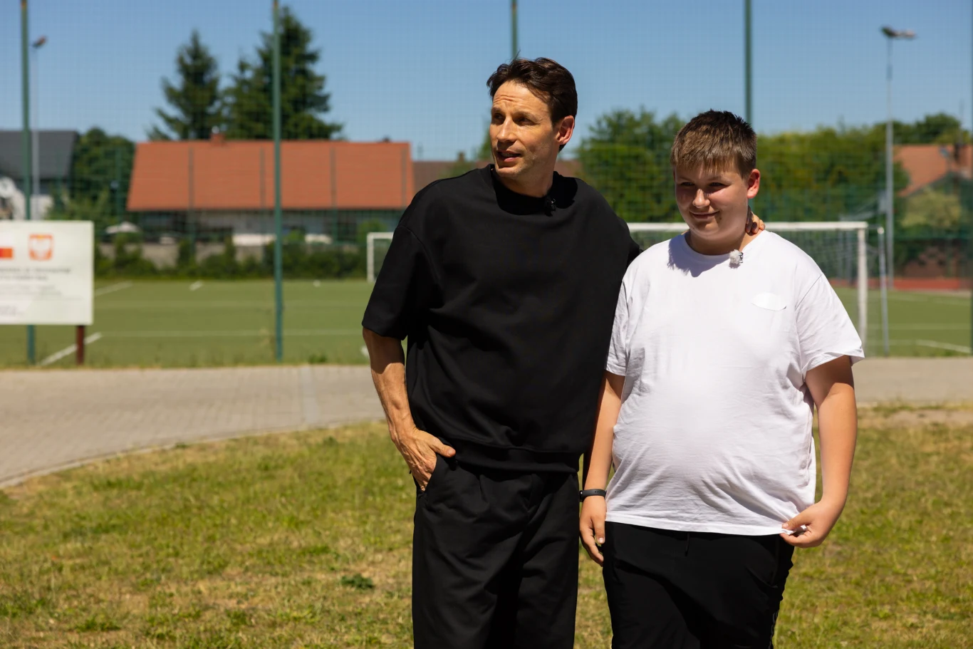 Nasz nowy dom Dwie osoby stoją na trawie przy boisku sportowym, jedna dorosła i jedno dziecko w jasnych i ciemnych ubraniach, w tle ogrodzenie i zabudowa mieszkalna.