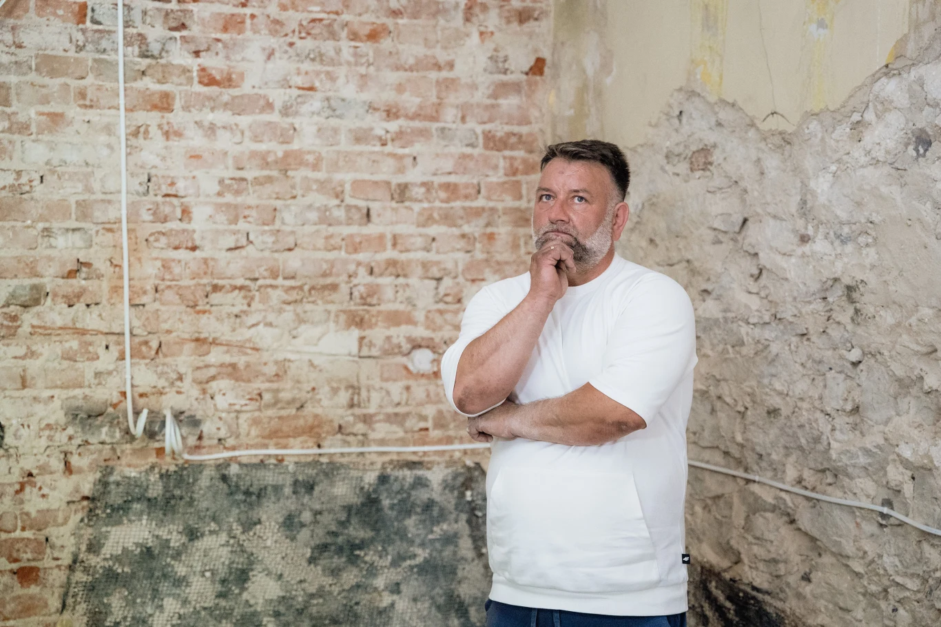 Nasz nowy dom Mężczyzna z brodą w białej koszulce w zamyśleniu opiera się o ścianę z odsłoniętej cegły i tynku, w tle widoczne przewody elektryczne i ślady remontu.