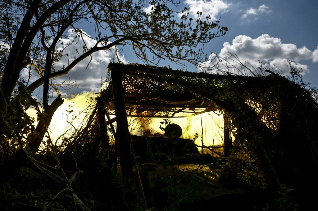 Ukraińscy żołnierze w obwodzie zaporoskim. Trwają walki o Pokrowsk i Hulajpole Ukryta pod kamuflażem armata polowa prowadzi ogień wśród gęstych zarośli, silne światło eksplozji rozświetla sylwetkę działa, a chmury na niebie podkreślają dramatyzm sceny.