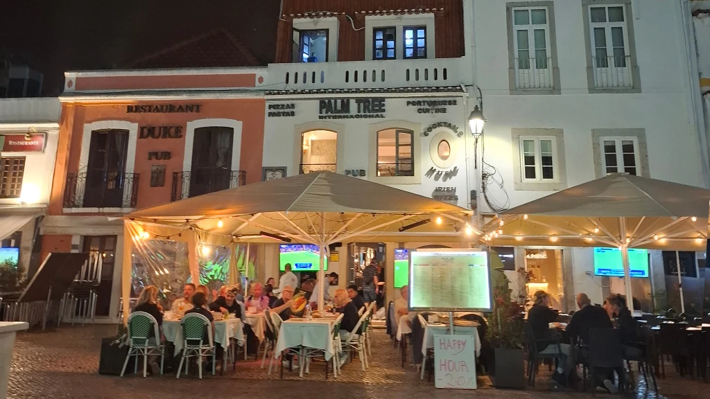 Cascais wieczorami tętni życiem Wieczorna scena na placu z jasnymi parasolami i ogródkiem restauracyjnym. Grupa osób siedzi przy stołach pod zadaszeniem, rozmawiając przy kolacji, otoczona delikatnym światłem lamp. W tle widoczne są elewacje budynków z szyldami restauracji i pubów.