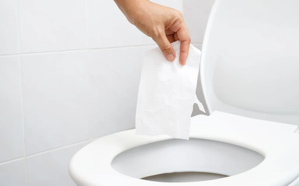 Zakup papieru to sposób na spuszczenie pieniędzy w toalecie? Japończycy znają rozwiązanie Ręka trzymająca papier toaletowy nad otwartą deską sedesu w jasnej łazience, ściany wyłożone białymi kafelkami.