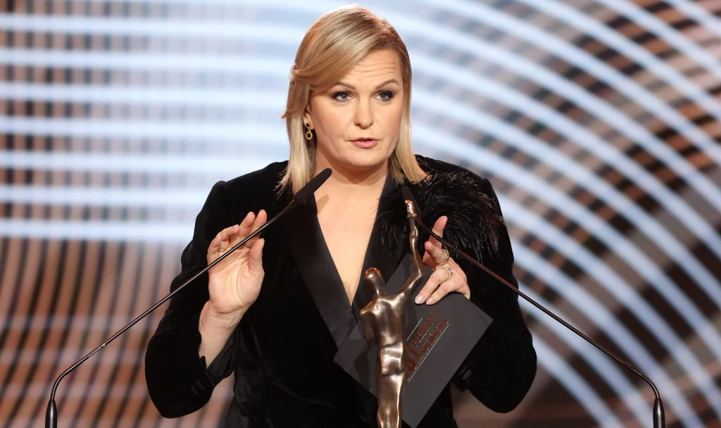 Otylia Jędrzejczak podczas tegorocznej Gali Mistrzów Sportu Kobieta w eleganckiej, czarnej sukni przemawia na scenie stojąc za mikrofonem i trzymając statuetkę w jednej dłoni oraz kopertę w drugiej, w tle widoczne geometryczne, jasne wzory.