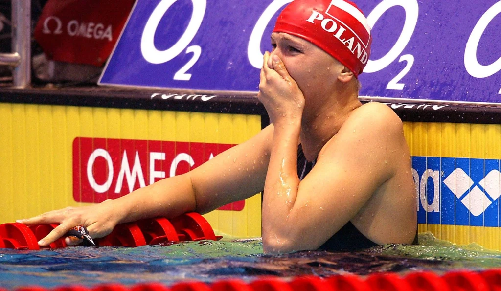 Otylia Jędrzejczak w czasach kariery zawodniczej. Łzy szczęścia na mecie rywalizacji Zawodniczka w czepku z napisem POLAND przytrzymuje się krawędzi basenu, zasłaniając usta dłonią, wyrażając silne emocje po wyścigu pływackim; w tle tablice wyników i logotypy sponsorów.