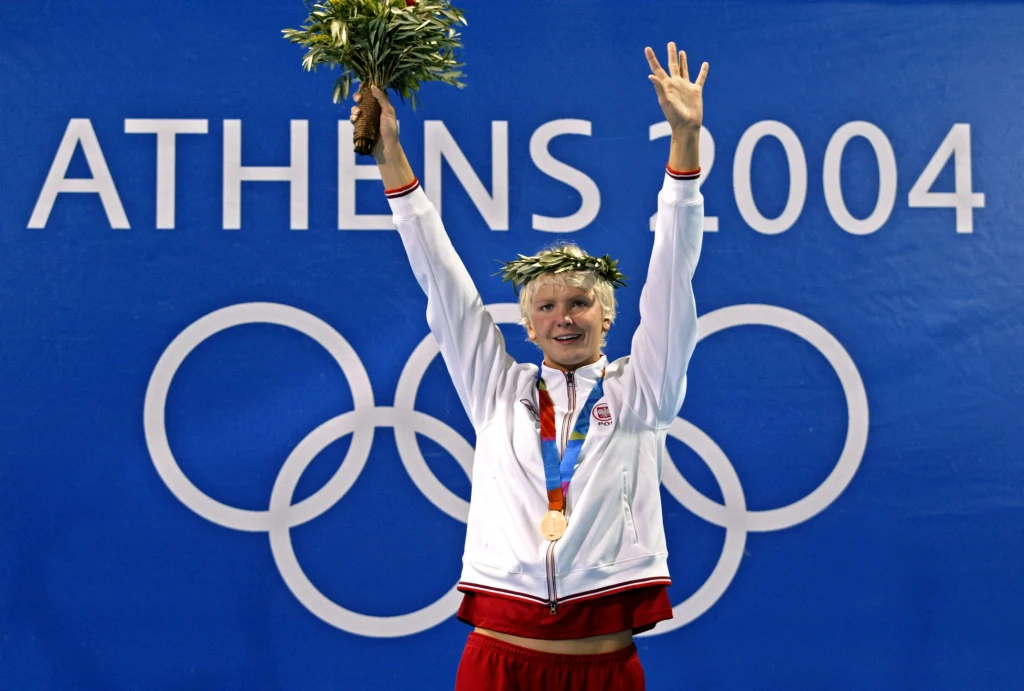 Otylia Jędrzejczak z olimpijskim złotem w Atenach Sportowiec w biało-czerwonym stroju z medalem olimpijskim i wieńcem laurowym na głowie unosi rękę w geście zwycięstwa na tle napisu Athens 2004 oraz olimpijskich kół.