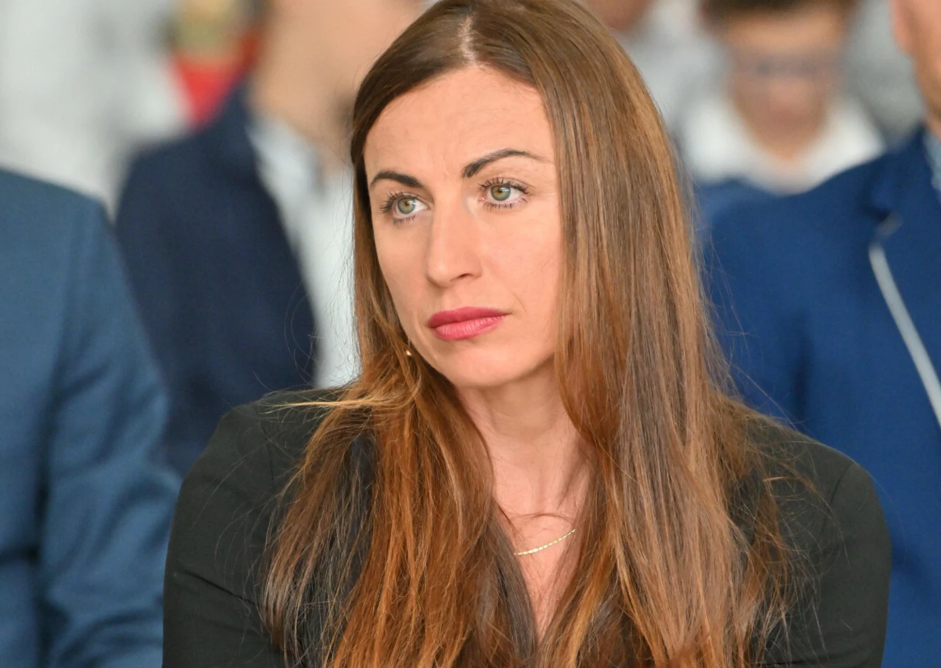 Kobieta o długich, brązowych włosach i jasnych oczach ubrana w czarną marynarkę siedzi w tłumie, skupiona, z delikatnym wyrazem zamyślenia na twarzy. W tle niewyraźne postacie innych osób.